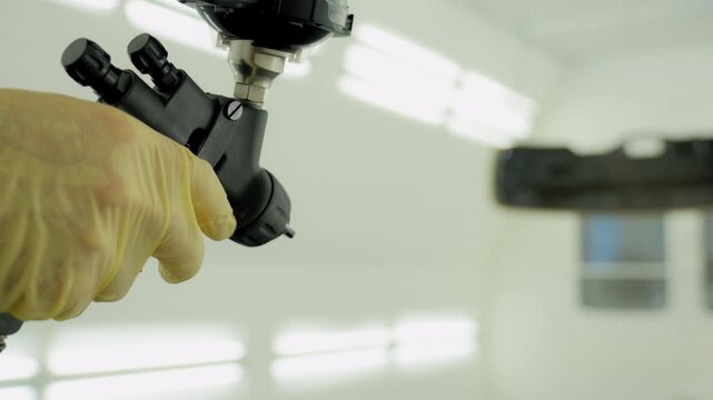 Professional auto technician applies black paint onto a small test panel using a spray gun inside a clean, well lit automotive paint booth. 