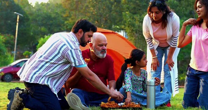 Indian family eating snacks while camping outdoors looking at camera, enjoying cheerful bonding moments, sharing laughter, relaxed conversations, warm interactions, and joyful quality time together