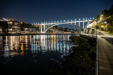 Obraz premium Brightly illuminated Arrabida Bridge and surrounding buildings reflected in calm water of Douro River in Porto, Portugal at night.