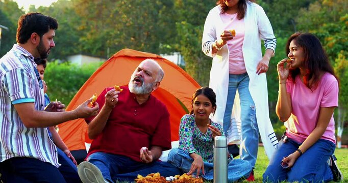 Indian family eating snacks while camping outdoors looking at camera, enjoying cheerful bonding moments, sharing laughter, relaxed conversations, warm interactions, and joyful quality time together