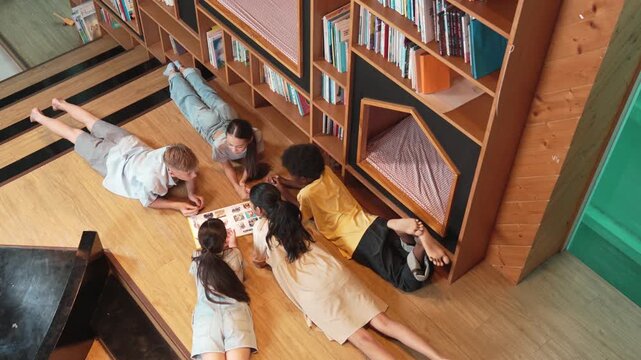 Group of diverse children lying down in circle while reading a book at library. Top view of girl sharing a magazine while pointing at interested topic and talking with lovely friends. Edification