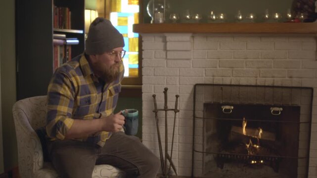 A man in plaid sits by the fireplace in a cozy fall living room setting, stirring and drinking tea. The vintage fireplace with logs on fire warms the living room. Shot in 60 frames for slow motion.