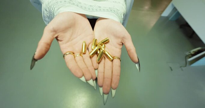 Close-up of a woman's hands with long black nails holding shiny golden pistol cartridges under studio light.