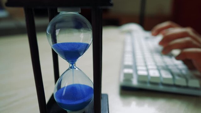 Close-up of an hourglass with blue sand next to a person typing on a keyboard - Powered by Adobe