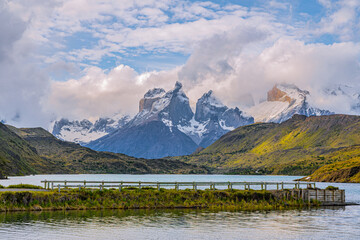 Dramatic mountain peaks rise above a calm lake and green hills in Torres del Paine National Park, Chile, under dynamic clouds and soft light.