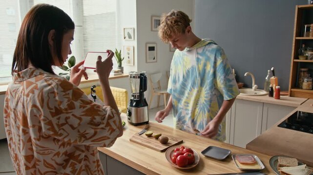 Caucasian teen girl holding smartphone in hands recording video of her male friend cooking at kitchen counter