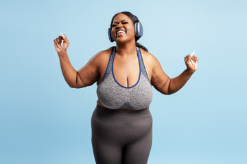 Happy, body positive Nigerian woman wearing stylish top listening music in headphones dancing