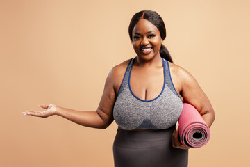 Smiling plus size model holding yoga amt showing with hand, copy space isolated on beige background