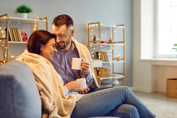 Fototapeta premium Happy cheerful joyful married couple enjoying good relationship, spending winter day off at home, cuddling on comfy cosy sofa in living room with Xmas lights, drinking hot tea, and having fun together