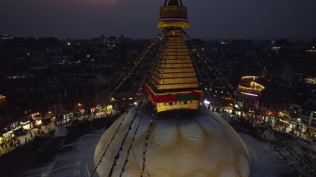 A calm night drone view of Boudhanath Stupa, with boudha and nath noted separately, glowing above Kathmandu. The sacred Buddhist landmark stands radiant in the quiet evening cityscape.