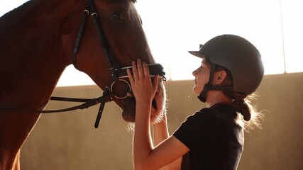 Woman wearing riding harness, adjusts horse's bridle in indoor arena, caring for beloved pet. Rider strokes horse with hand, owner taking care of animal. Concept: equestrian sport, riding school