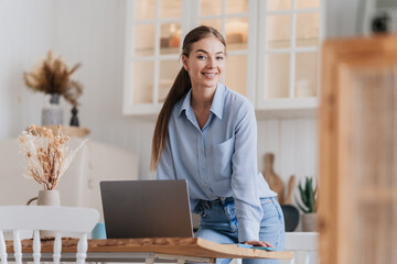 A smiling woman stands with confidence next to her laptop in a bright kitchen, radiating positivity and readiness.