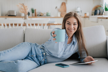 A cheerful woman in a light blue blouse lies on a sofa, raising a mug towards the camera, next to her smartphone and tablet, exuding a relaxed vibe.