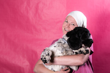 Portrait of a woman wearing a headscarf associated with cancer treatment, tenderly hugging her Cocker Spaniel dog against a pink background. Image conveying emotional support, pet companionship