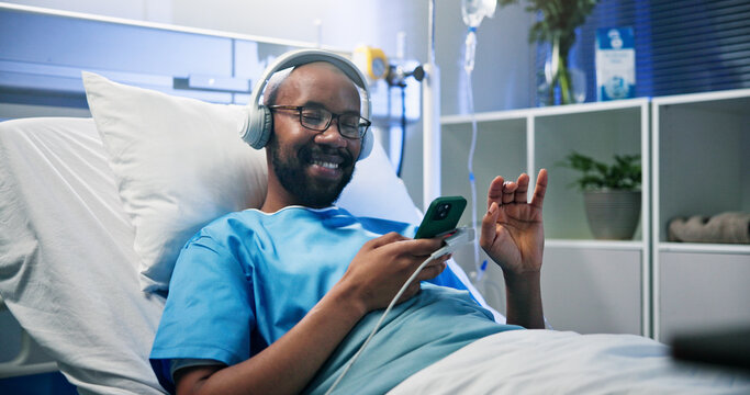 Music, phone and smile of black man in hospital bed for medical recovery, streaming or treatment. App, audio and headphones with happy patient lying in healthcare clinic for rehabilitation or rest - Powered by Adobe