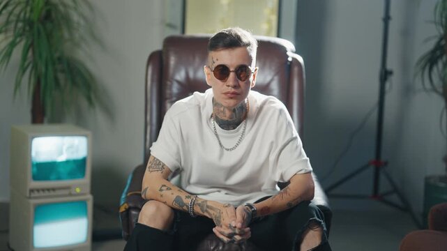 Portrait of strange man with tattoos on face and body sitting alone in studio. Trendy and stylish male person looking at camera through black sunglasses, contemporary music and fashion, hip-hop singer