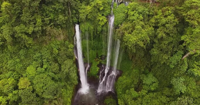 Drone video captures a stunning aerial view of a multi-stream waterfall cascading through lush jungle foliage near Gitgit, Bali. No people visible, vibrant greens dominate the scene.