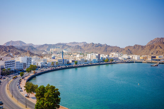 A stunning panoramic view of Muttrah Corniche in Muscat, Oman, featuring turquoise blue water, a curving coastal road, white traditional buildings, and rugged desert mountains rising behind the city