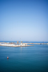 A wide coastal view of Muscat Harbor in Oman, featuring calm blue water, a breakwater barrier stretching across the horizon, port facilities, cranes, and a distinctive control tower