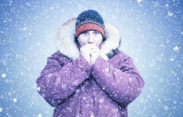 Freezing man in a winter jacket warms his hands in heavy snowfall. Cold weather, blizzard conditions and harsh winter survival concept on an icy outdoor background.