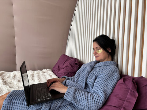 Mature brunette woman with under-eye patches working on a laptop at home, communicating online and freelancing while combining self-care, calm atmosphere, and remote work