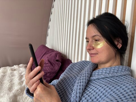 Mature brunette woman with under-eye patches working on a smartphone at home, chatting and freelancing while relaxing and combining skincare with remote work.