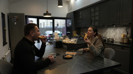 A couple in the kitchen is eating buns and drinking tea.