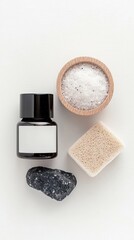 A flat lay composition of spa and wellness items including a black stone, bath salts in a wooden bowl, a pumice stone, and a dark bottle with a blank label.