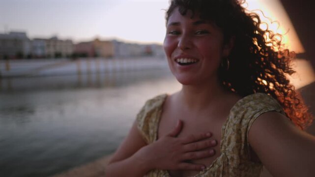 Beautiful curly haired woman smiling and recording a selfie video call from a city riverside at sunset, happily waving, gesturing, and blowing a kiss to the camera with lens flare