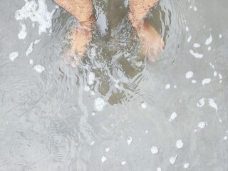 A low-angle shot shows bare feet partially submerged in shallow, foamy ocean water that washes over dark sand, creating gentle ripples.
