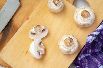 Arrangement of whole and sliced button mushrooms on a wooden cutting board, with a purple cloth and knife in the background. Overhead shot for cooking and kitchen concepts.