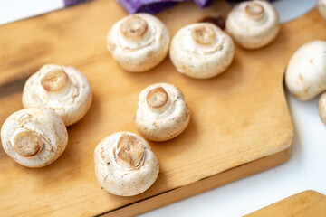 Arrangement of whole and sliced button mushrooms on a wooden cutting board, with a purple cloth and knife in the background. Overhead shot for cooking and kitchen concepts.