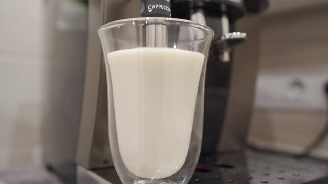 A glass cup filled with milk sits below the metal milk frothing apparatus of an espresso machine. The machine prepares for the frothing process, activating its pump
