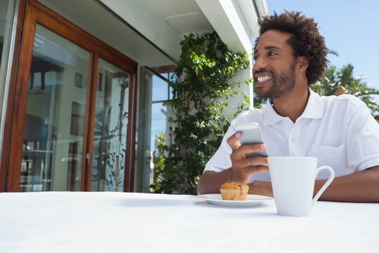 African American midadult man sitting on sunny patio using phone beside mug and muffin, copy space