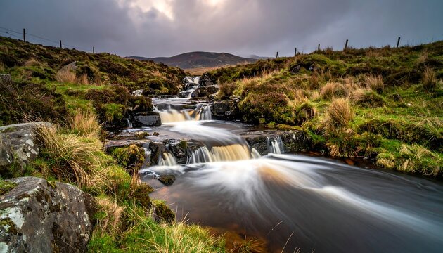 A winding stream cascades over rocks amidst green vegetation under a cloudy sky, a distant mountain visible