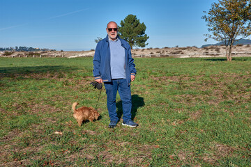 Elderly man walking along a rural path with his dog on a sunny day in a green setting with trees and a cityscape in the background