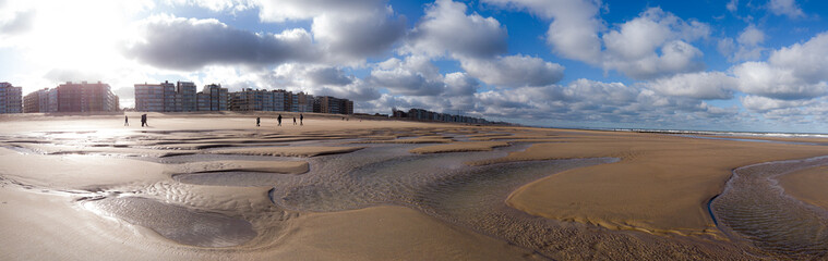 A wide, sunlit beach with shallow tidal pools and rippled wet sand stretches toward a row of urban buildings along the coast. A few figures walk the shoreline beneath a dramatic, cloud-filled sky.