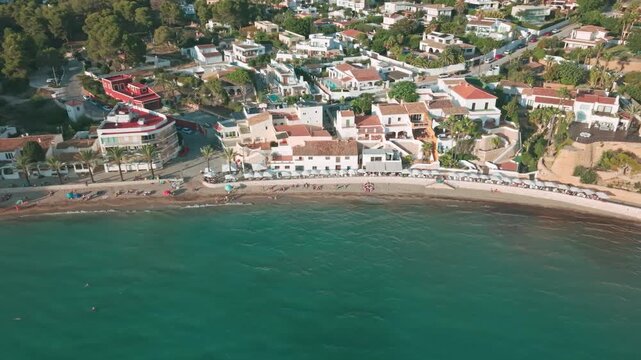 Aerial drone footage of Moraira town and beach on the Costa Blanca, Spain. Scenic coastal view with Mediterranean water, houses, and palm-lined promenade.