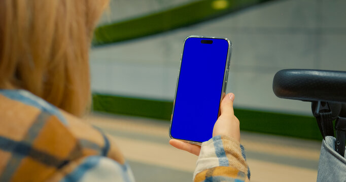 Young woman on subway platform, holding smartphone with blue screen mockup, representing urban digital connectivity and modern commuting lifestyle. Blue screen chroma key.