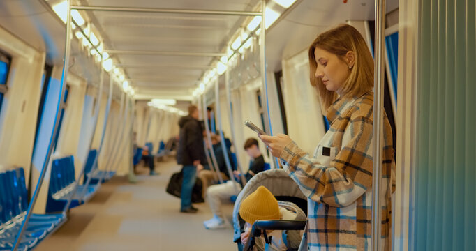 Young mother multitasking, using smartphone while pushing baby stroller inside busy subway carriage, embodying digital parenting lifestyle