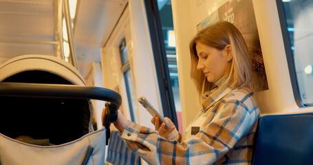Young woman commuting by subway, sitting near baby stroller and browsing smartphone while navigating urban public transport experience