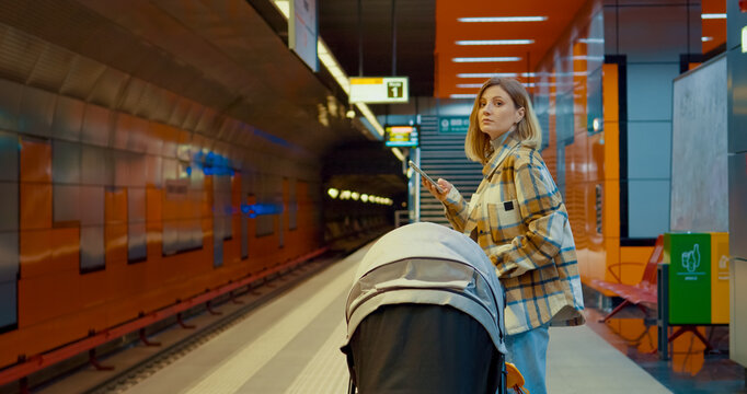 Woman waiting on subway platform, holding smartphone while pushing baby stroller, capturing urban parenting moment in modern transportation setting - Powered by Adobe