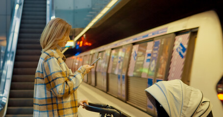 Multitasking female waiting at subway platform, holding baby stroller while checking smartphone during urban commute through modern city transportation hub