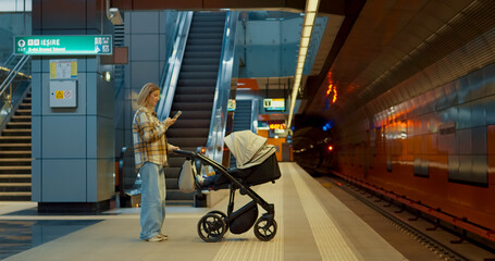 Young mother multitasking, checking smartphone while waiting with baby stroller at subway station...