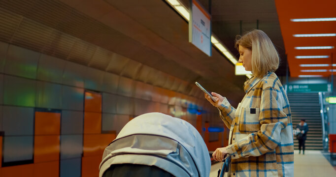 Multitasking parent navigating urban commute, using smartphone while standing with baby stroller at metro platform
