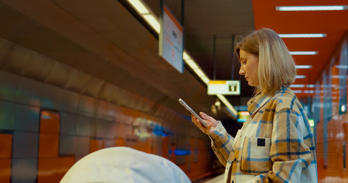Woman navigating urban commute, using smartphone while standing with baby stroller at metro platform