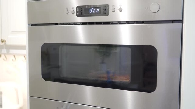 Stainless steel and glass built-in microwave oven. It is actively heating food, evidenced by the digital countdown and warm interior glow. The process generates a steady whirring sound