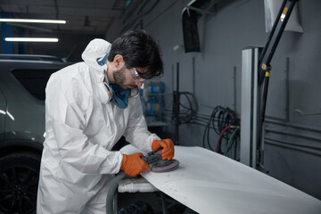An auto body technician in a protective suit meticulously sands a car hood at a repair shop