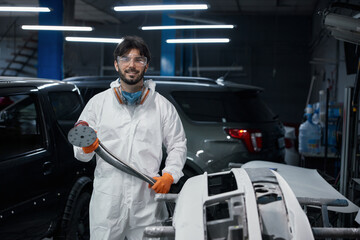An auto body technician sands a car bumper in a paint shop, wearing safety gear