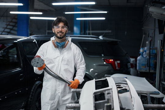 An auto body technician sands a car bumper in a paint shop, wearing safety gear